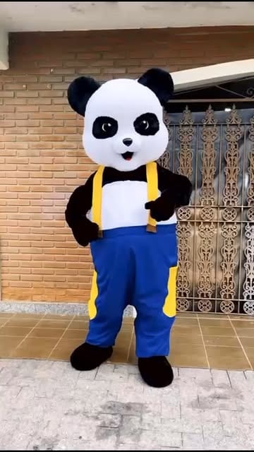 Mascote de panda com macacão azul e suspensórios amarelos, de corpo inteiro na calçada