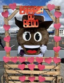 Mascote Pinhão, marrom e de chapéu de caubói, na barraca do beijo enfeitada com corações numa festa junina