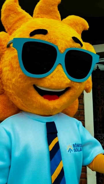 Mascote de sol amarelo e peludo com óculos escuros azuis, camisa azul-clara e gravata listrada